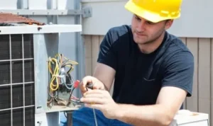 an electrician wearing a yellow hard hat repairing the electrical wiring on an outdoors HVAC system